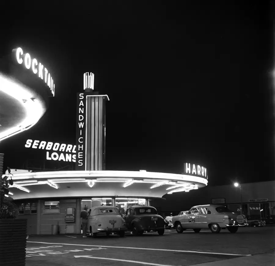 Black-and-white photo of a retro diner at night with neon signs reading “Cocktails,” “Sandwiches,” and “Seaboard Loans.” Vintage cars are parked outside beneath the glowing lights.