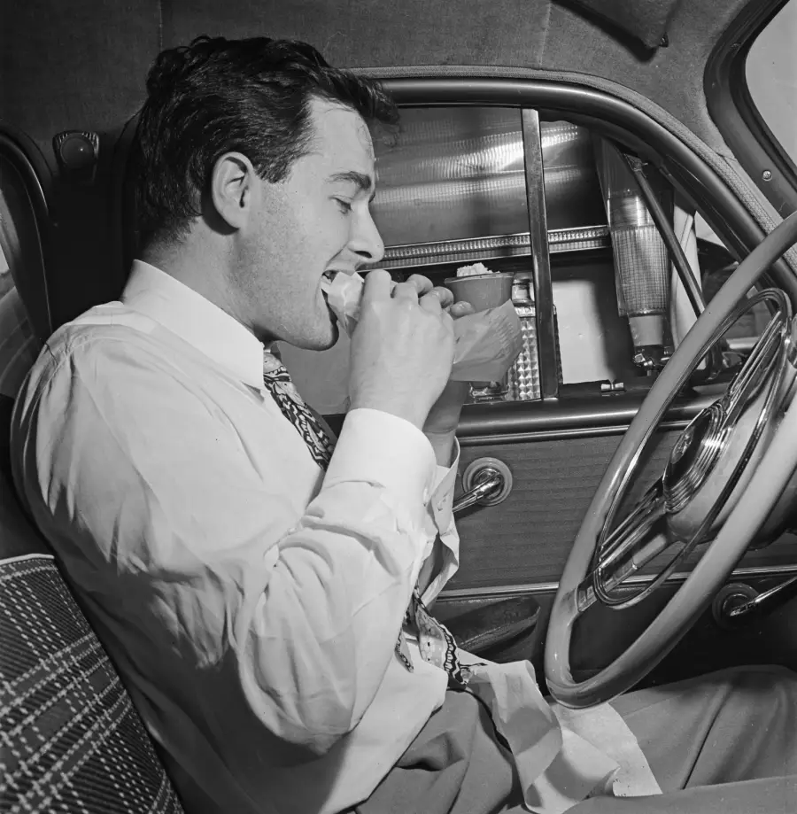 A man in a dress shirt and tie sits in the driver’s seat of a car, eating a sandwich. He appears to be enjoying his meal, and the car’s interior is visible, including the steering wheel and patterned seat.