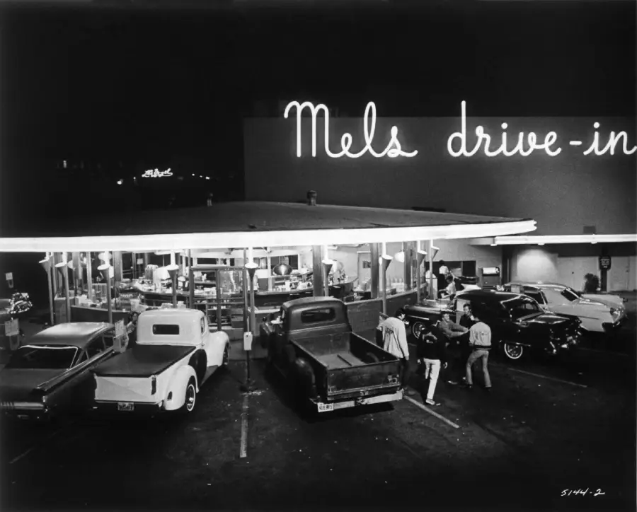 Black and white photo of Mel’s Drive-In at night, with several vintage cars parked and people gathered under a bright neon sign, evoking a classic 1950s diner scene.