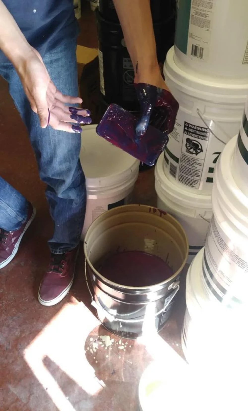 A person with purple paint on their hands and a cup stands next to several white buckets, with an open bucket of purple paint on the floor.