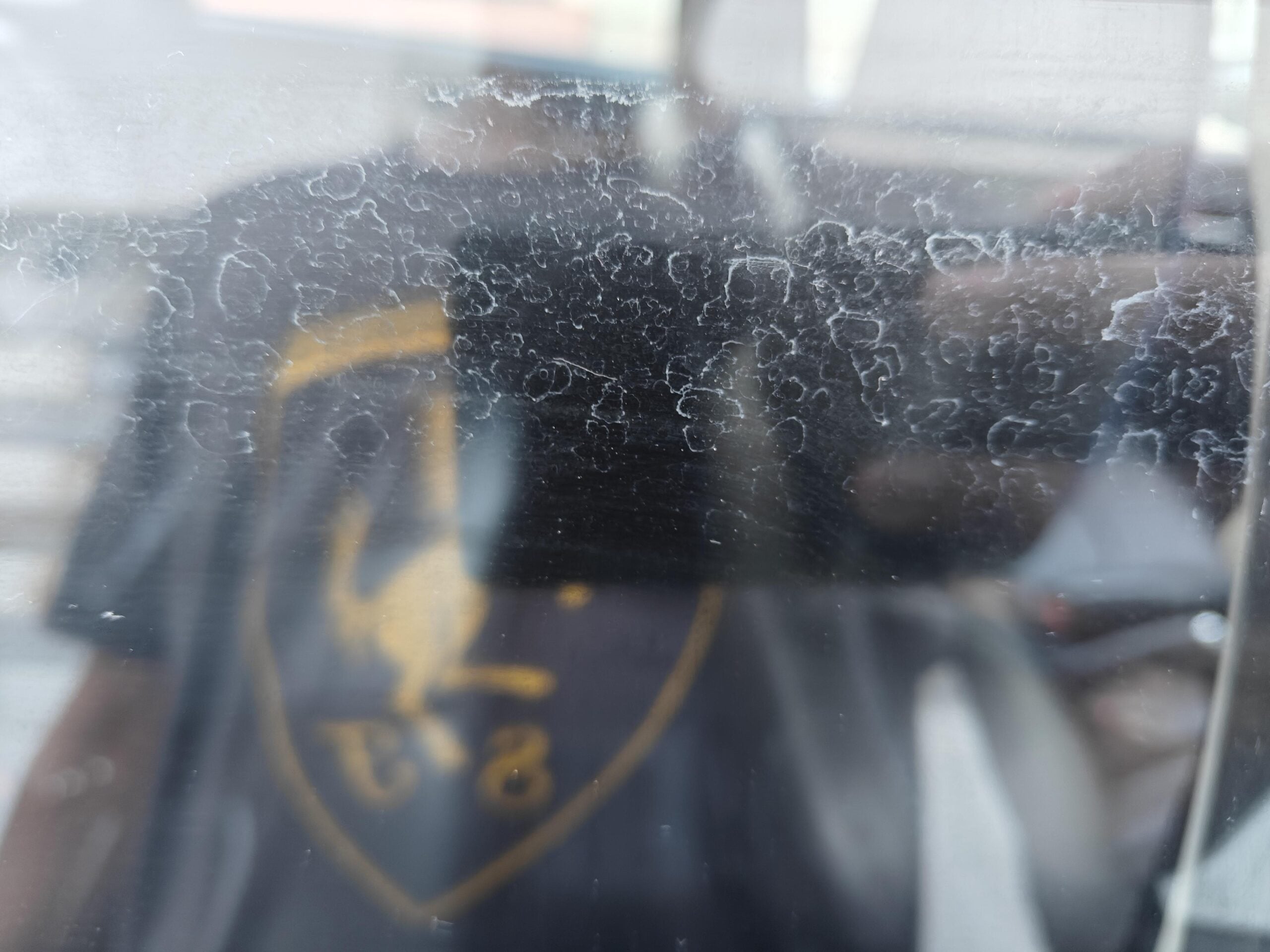 Blurry reflection of a person wearing a dark shirt with a yellow logo, seen through a glass surface with white streaks and spots.