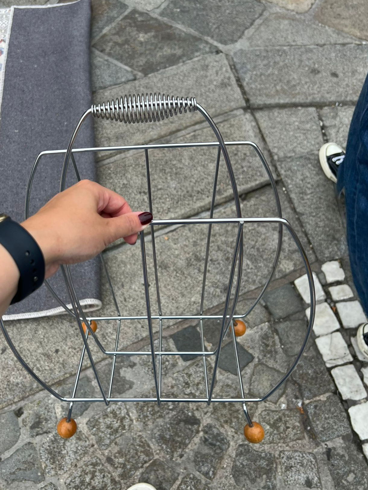 A hand holding a silver wire magazine rack with wooden feet, positioned on a stone pavement with parts of two people’s shoes visible.