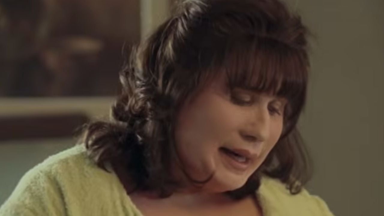 A woman with brown hair and bangs, wearing a light green top, looks down with a gentle expression in a softly lit room.