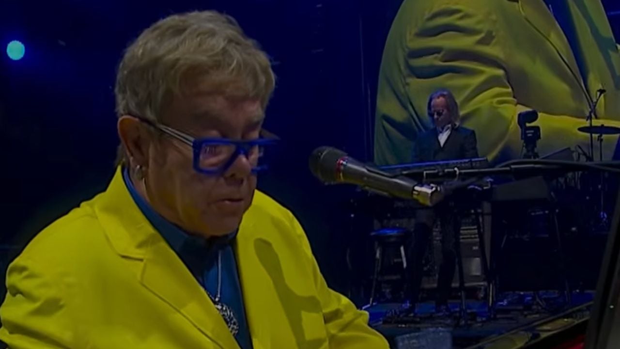 A person in a bright yellow jacket and blue glasses plays piano on stage, with another musician and a large screen in the background.