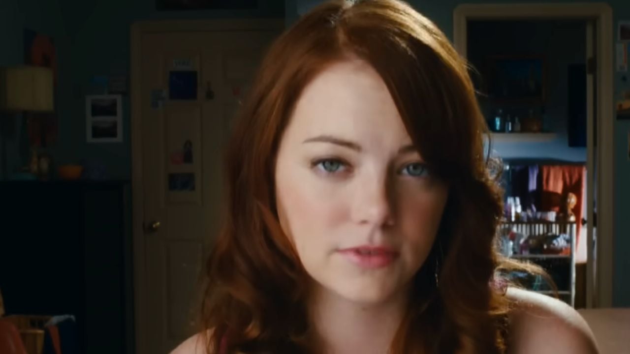 A young woman with long red hair and light eyes looks into the camera, slightly smiling, in a dimly lit bedroom with a closed door and shelves in the background.