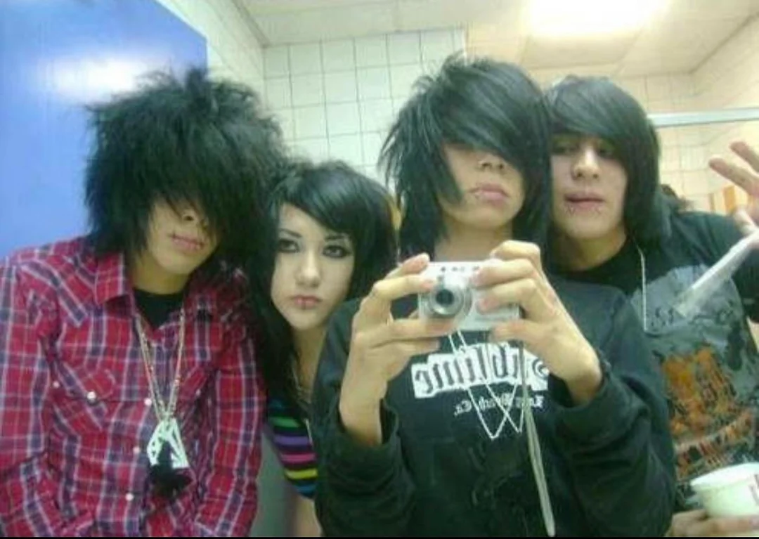 Four teens with emo hairstyles pose for a selfie in a bathroom mirror. Three have long black hair covering their faces; one wears a plaid shirt and another wears black with chains. The background shows tiled walls and stalls.