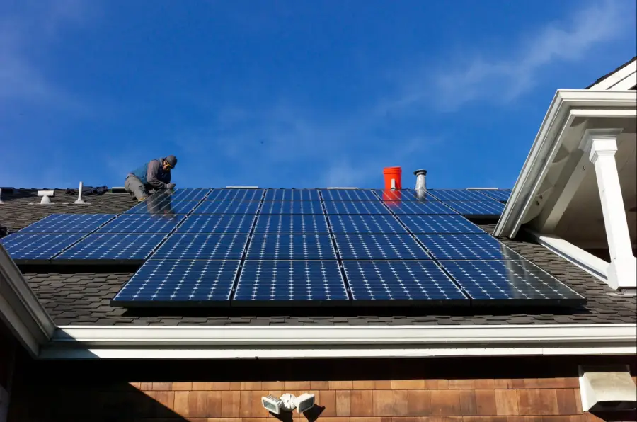 A person in work clothes installs solar panels on the roof of a house under a clear blue sky.