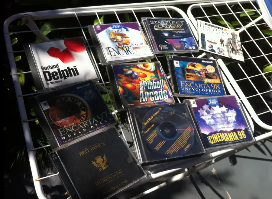 A collection of old PC CD-ROMs, including Encarta encyclopedias, Star Wars Jedi Academy, Pinball Arcade, Britannic Encyclopedia, and Borland Delphi, displayed on a white metal rack outdoors in sunlight.