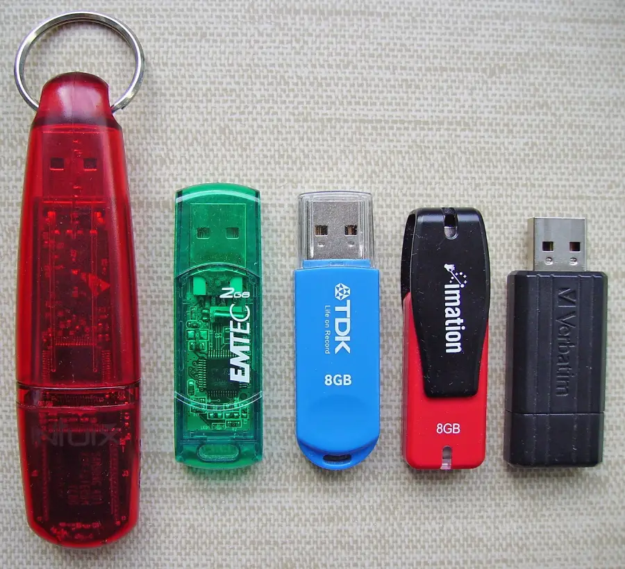 Five USB flash drives of various colors and brands are arranged vertically on a beige fabric background. The drives are red, green, blue, black with red, and black.