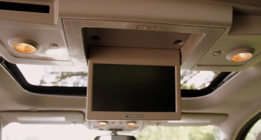 A Panasonic flip-down screen is mounted on the ceiling of a vehicle, with overhead lights and a sunroof visible in the background.