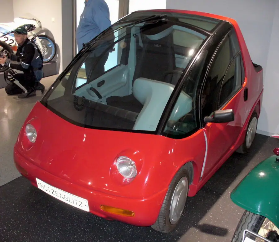A small, red, two-seater car with a rounded front, large windshield, and compact design is on display indoors. A person and a motorcycle are visible in the background.