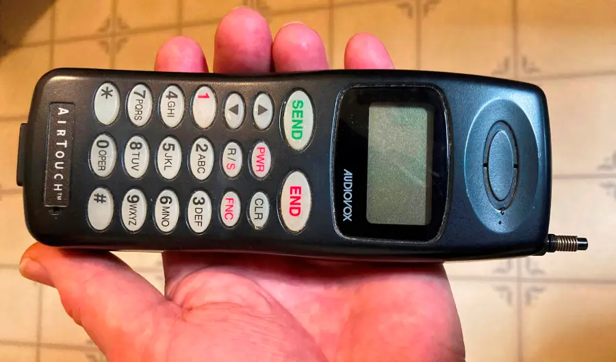 A person holding an old-fashioned Audiovox cell phone with large buttons and a small screen. The phone’s antenna is extended, and the background is a tiled floor.