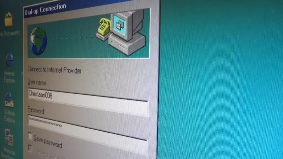 Close-up of a Windows 98 dial-up connection window showing fields for username and password, with an old computer and phone icon, over a teal desktop background.