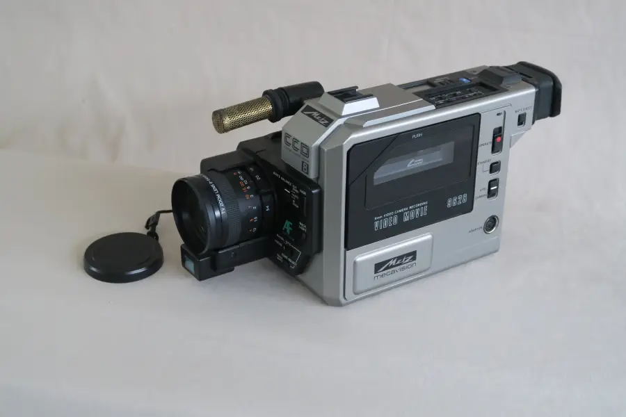 A vintage silver video camera with a black lens, microphone, and viewfinder sits on a light surface. The lens cap is off and lies beside the camera.
