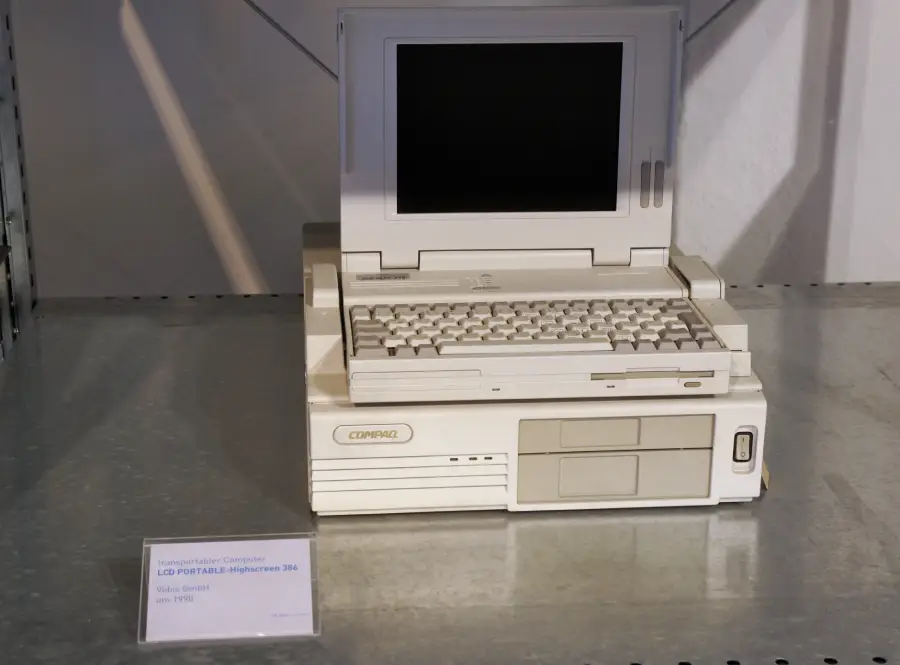 A vintage Compaq laptop with a monochrome screen and keyboard sits on a shelf. An information plaque is displayed to the left of the computer.