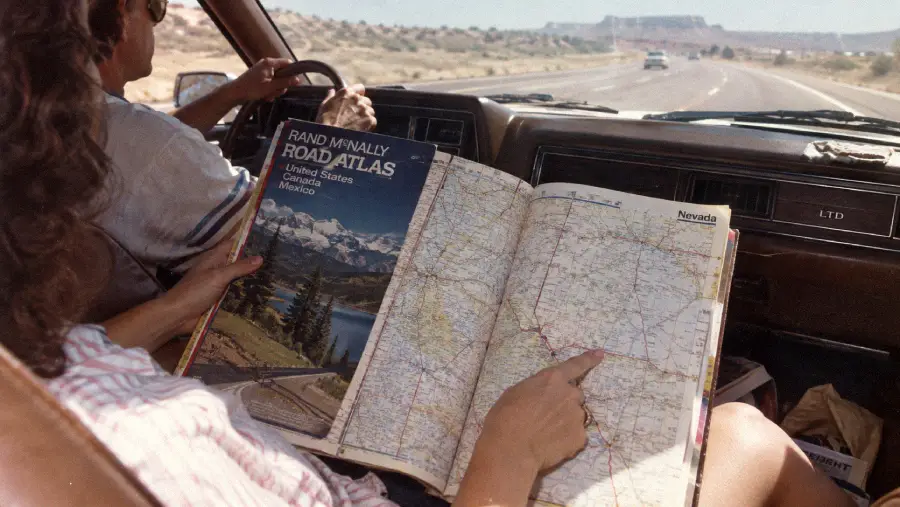Two people are driving on a highway in a car. The passenger holds a large Rand McNally road atlas and points at a map of Nevada, while the driver keeps one hand on the steering wheel. The road stretches into the desert landscape.