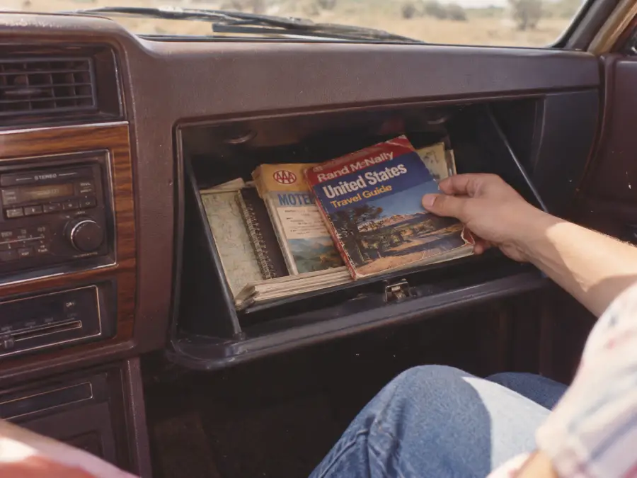 A person sitting in a car opens the glove compartment and pulls out a Rand McNally United States travel guide, with maps and other booklets also visible inside.