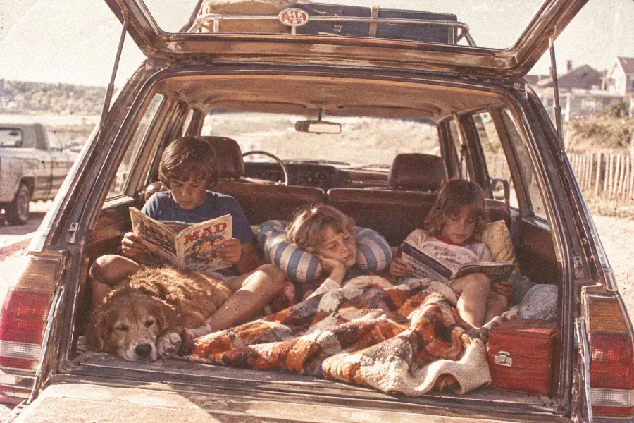 Three children read magazines in the back of a station wagon with a dog, a blanket, and a cooler. The car’s hatch is open, and the scene appears to be at a beach or a vacation spot.