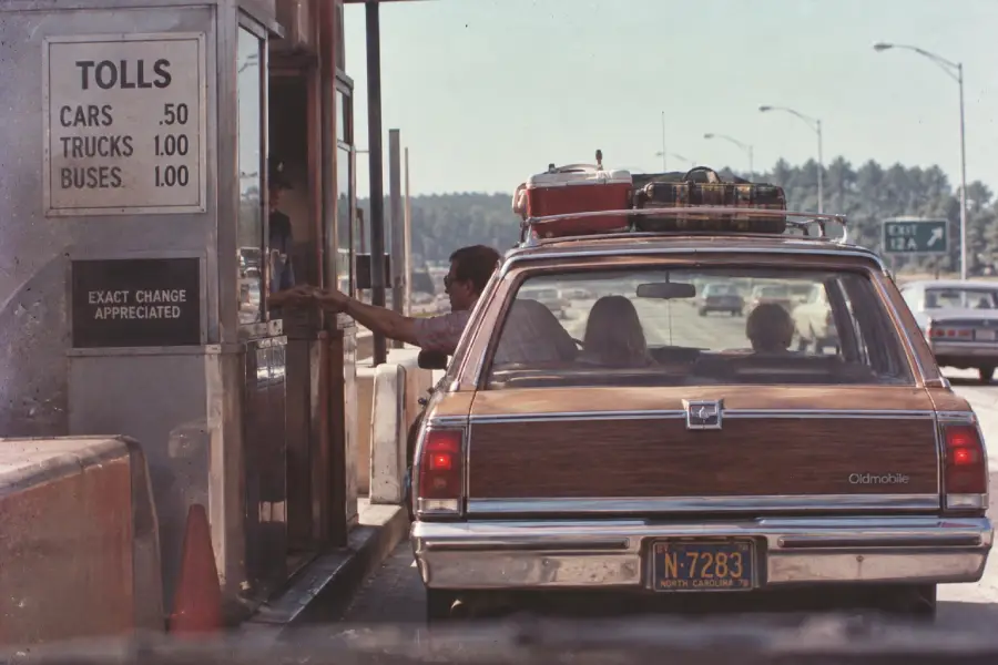 A station wagon with luggage on top stops at a toll booth. A person hands money to the attendant. The toll sign lists prices for cars, trucks, and buses. The car has a North Carolina license plate.