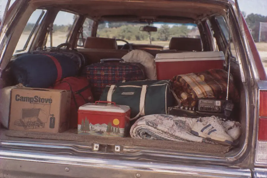 The open trunk of a station wagon packed with camping gear, bags, coolers, a box labeled “CampStove,” blankets, a radio, rolled sleeping bags, and a pair of white sneakers.