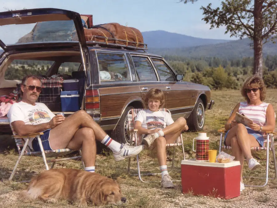 A family relaxes on lawn chairs by a station wagon with luggage on top. A dog lies nearby, and there’s a red cooler and picnic items in front of them. Trees and mountains are in the background.
