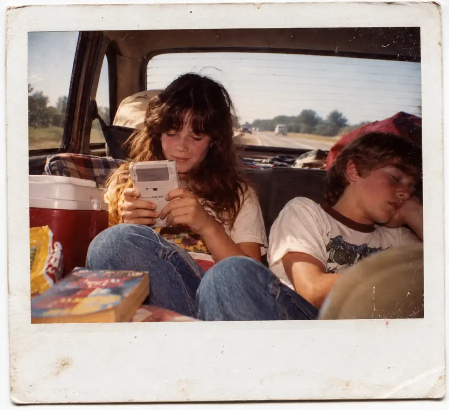 A girl plays a Game Boy while sitting in the back of a car, and a boy next to her sleeps. Sunlight streams in through the rear window, and books and snacks are nearby. It appears to be a road trip.