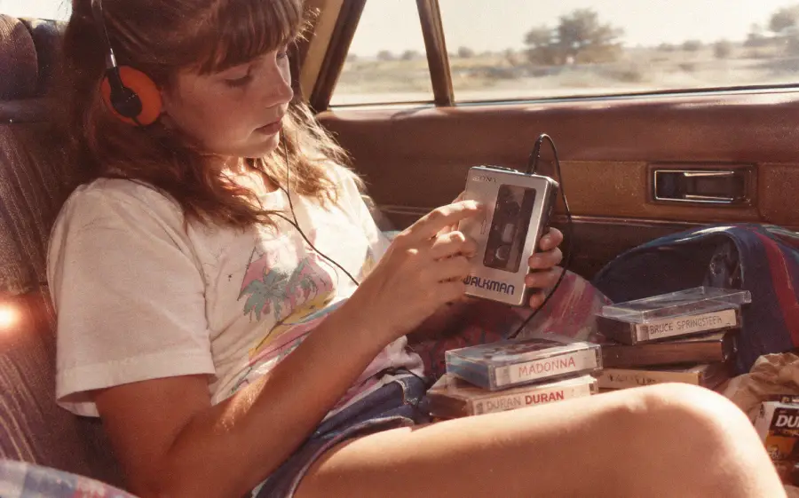 A girl sits in a car wearing headphones, holding a Sony Walkman. Several cassette tapes, including ones labeled “Madonna” and “Duran Duran,” are scattered on her lap and nearby. Sunlight streams in through the window.