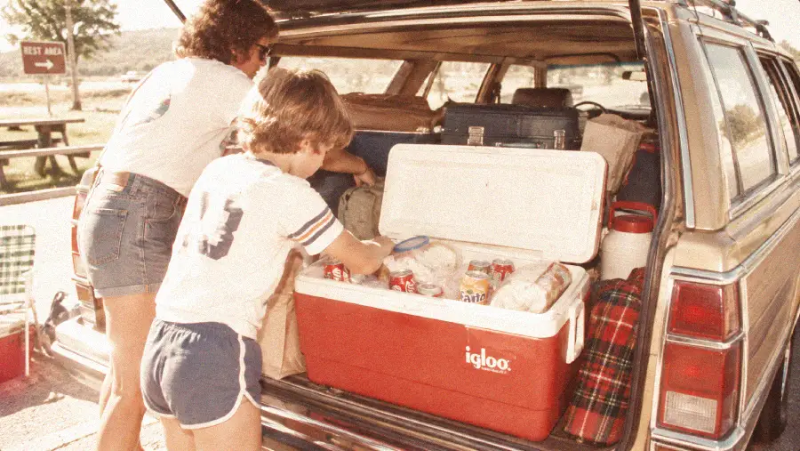 A child and an adult load a red Igloo cooler filled with drinks and food into the back of a station wagon packed for a road trip.