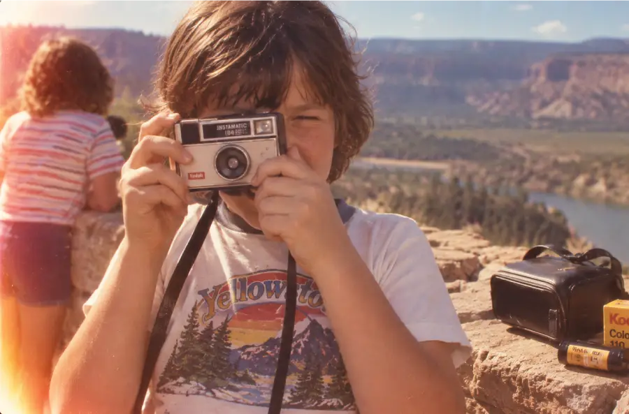 A child holding a vintage camera stands outdoors at a scenic overlook. Another child in striped clothes is in the background. A camera case and Kodak film box sit nearby. Mountains and river visible in the distance.