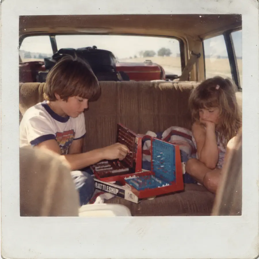 Two children sit in the backseat of a car playing the board game Battleship. Luggage is piled in the back, and scenery is visible through the windows, suggesting a road trip.