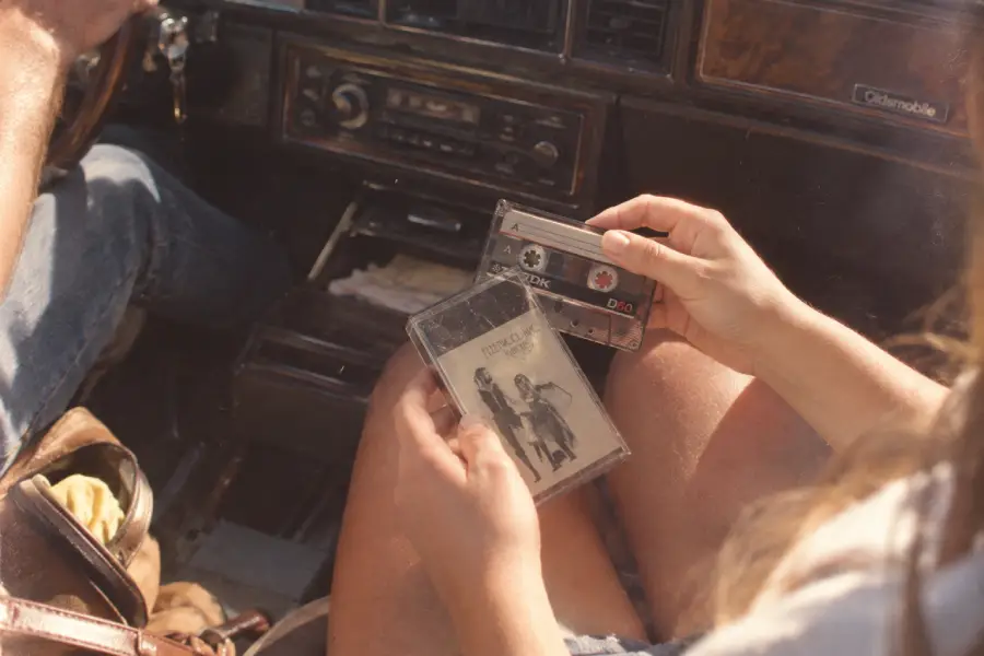 A person sitting in a car holds two cassette tapes, one labeled TDK and another in a clear case with an illustrated cover, next to a dashboard with a cassette player.