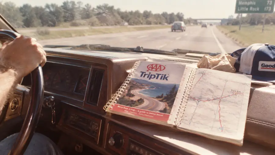 A person drives a car on a highway. On the dashboard are a AAA TripTik map, a crumpled paper bag, a trucker hat, and the car radio. A green road sign shows directions to Memphis and Little Rock.