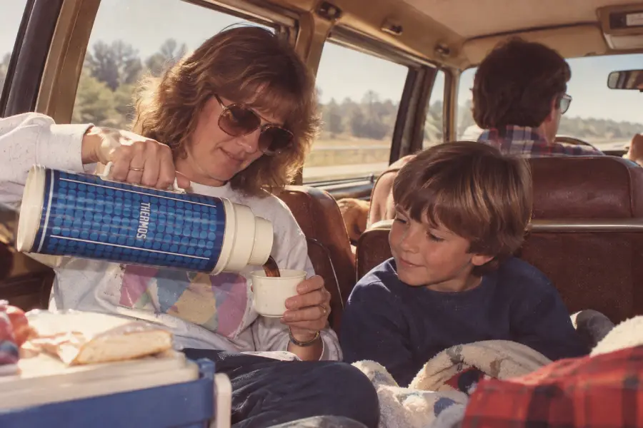 A woman pours a drink from a blue Thermos for a smiling young boy inside a car. They sit together with a blanket, and two other people are seated in front of them. Sunlight streams through the windows.