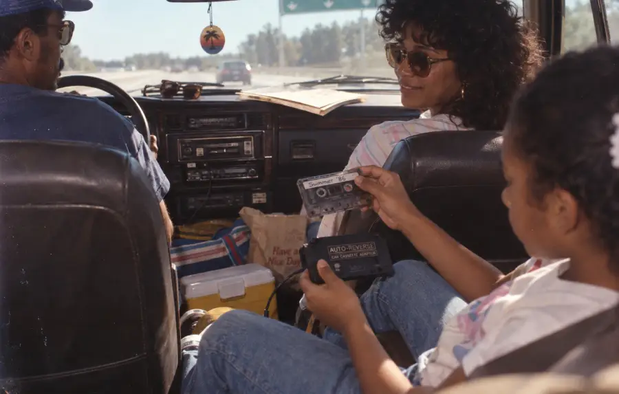 Two people sit in the front seats of a car while a passenger in the back holds a cassette tape and a tape case. The car is driving on a highway, and sunlight is coming through the windows.