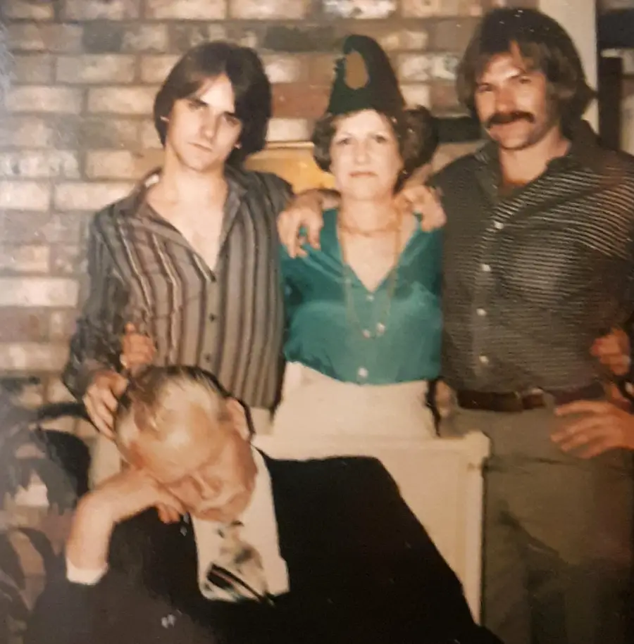 Three people stand in front of a brick wall, posing together with their arms around each other, while an older man sits in front of them, appearing to be asleep.