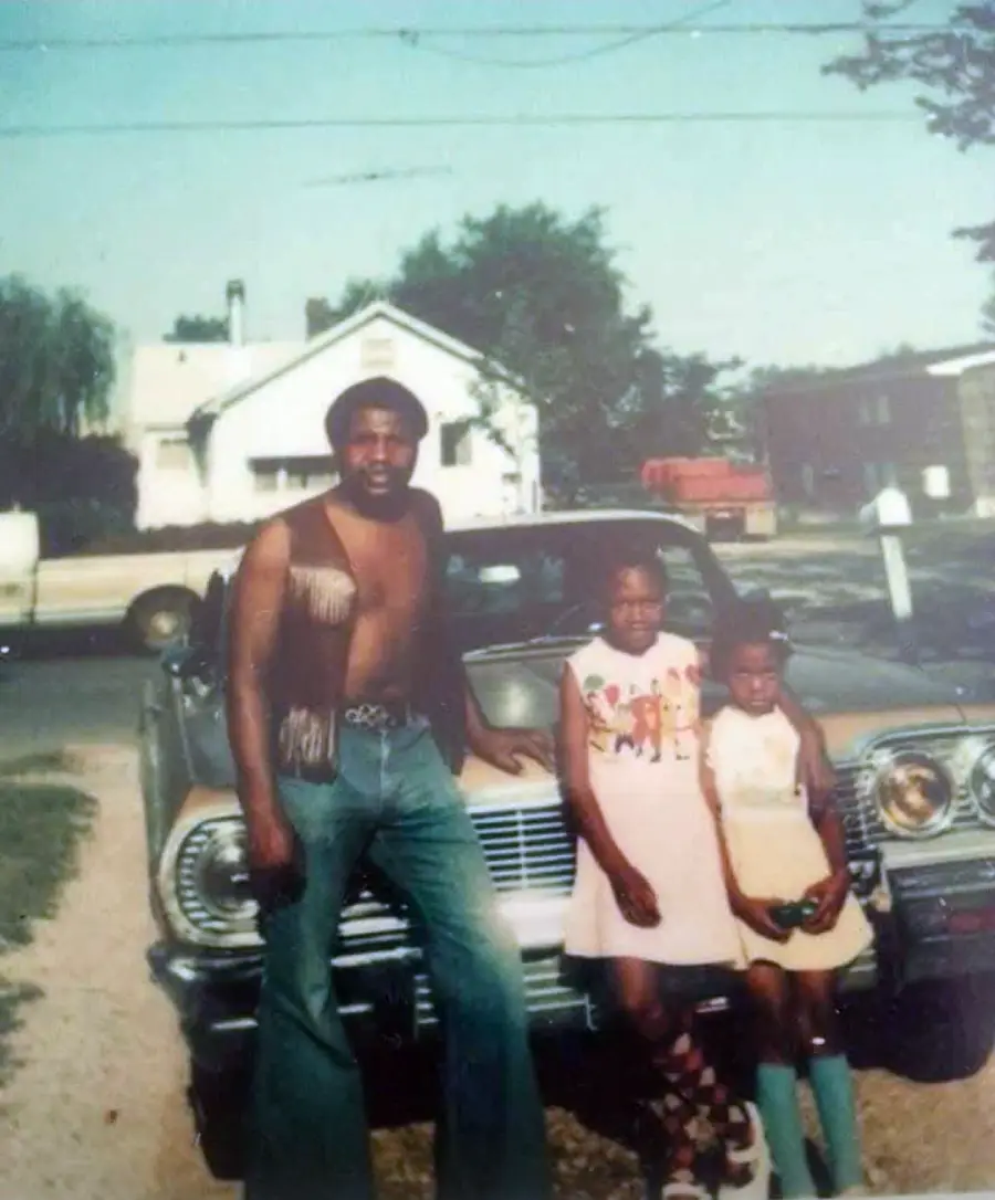 A man in a fringed vest stands beside two girls in dresses in front of a classic car, with houses, trees, and a mailbox visible in the background.