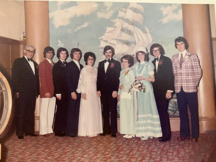 A wedding party from the 1970s stands indoors in front of a mural of a ship, wearing formal suits and dresses, smiling at the camera.