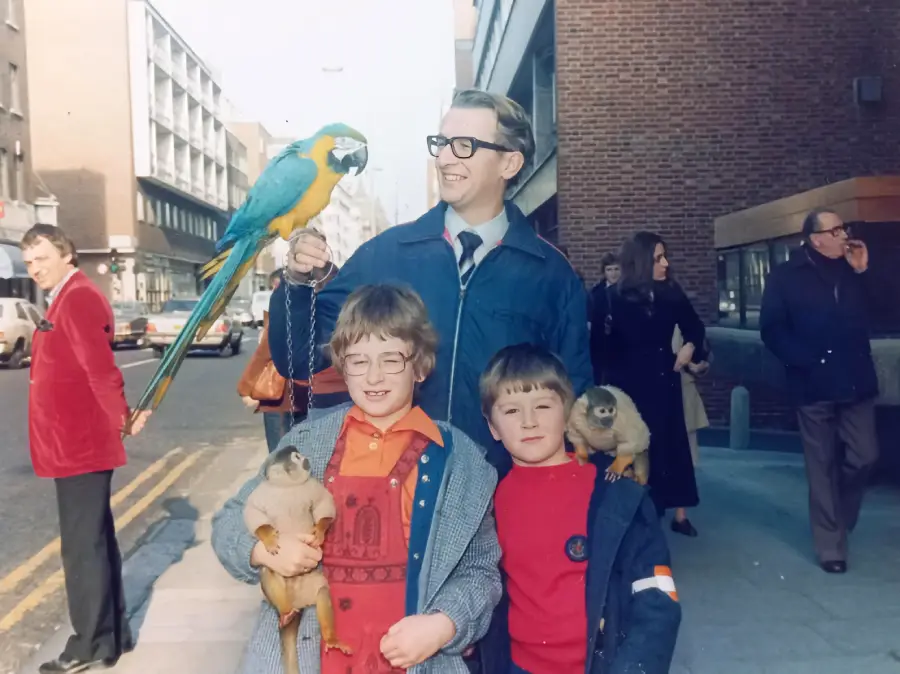 A man and two children pose on a city street holding monkeys, with a blue-and-yellow parrot perched on the man’s hand. Other people walk and stand nearby. It appears to be daytime.