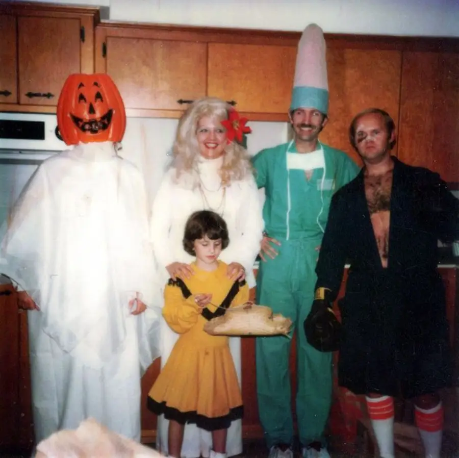 Five people in costumes stand in a kitchen: a ghost with a pumpkin head, a woman in white, a man in green with a tall pink hat, a child in yellow, and a man in boxing gear with a robe and gloves.