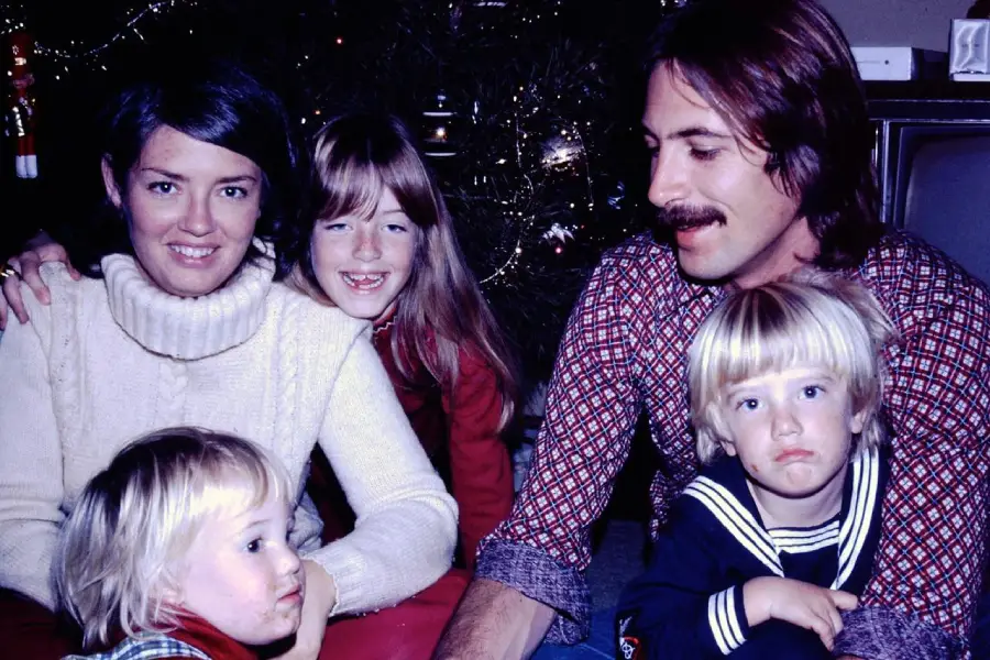 A family of five poses by a Christmas tree. Two adults and three children smile, except for one boy who looks sad. The adults wear sweaters, and the tree has lights and ornaments in the background.