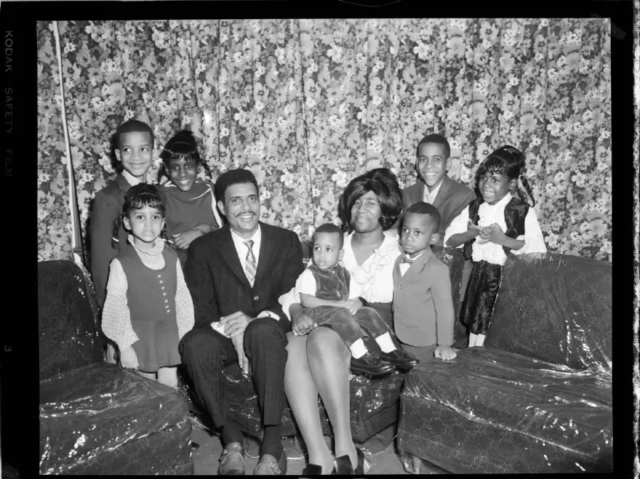 A family of nine, dressed formally, poses for a photo in a living room with floral curtains. Three adults and six children sit and stand together on plastic-covered furniture, smiling at the camera.