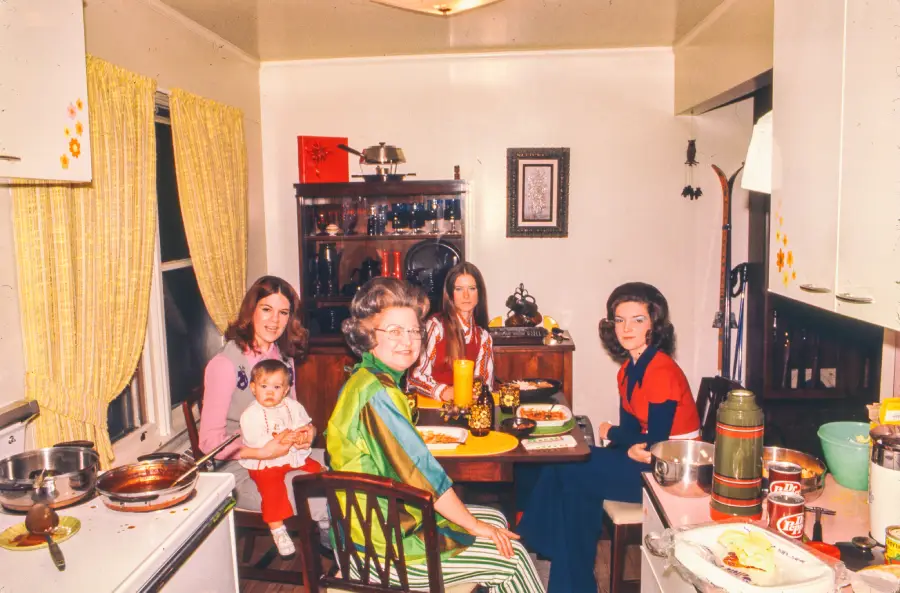 Four women and a baby sit at a small kitchen table set with plates of food, drinks, and condiments. Bright yellow curtains are on the windows. Kitchen utensils and dishes are visible on counters and shelves.