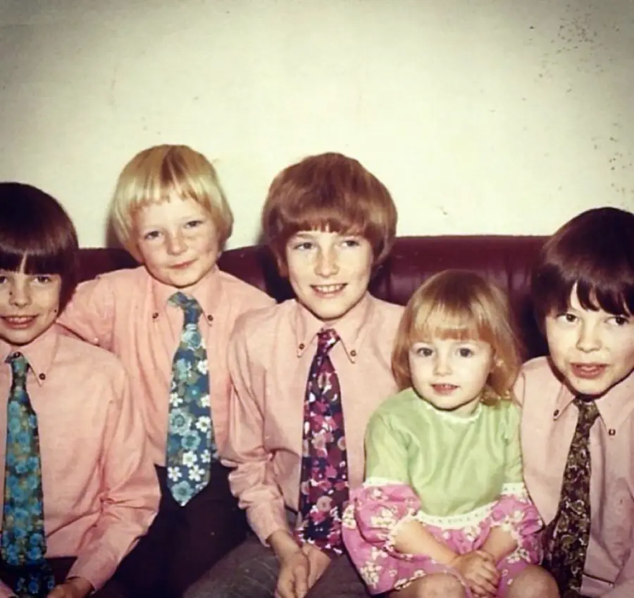 Five children sit on a couch. Four boys wear pink shirts and patterned ties; a little girl in a green and pink dress sits in the middle, smiling. The background is a plain wall.