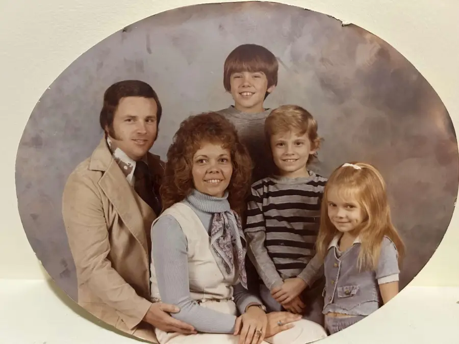 A vintage family portrait of two adults and three children posing together, all smiling. The adults are seated in front, while the children stand and sit around them, against a neutral gray background.