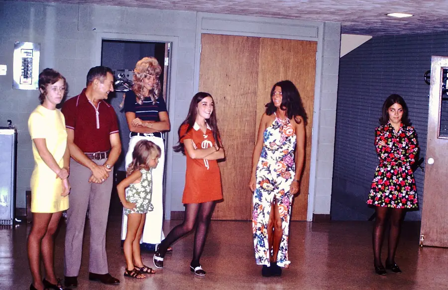A group of six people, including children and adults, stand indoors wearing colorful 1970s outfits, with a wooden door and water fountain in the background.