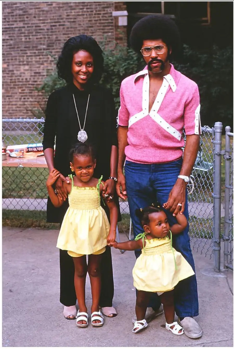 A smiling family of four poses outdoors. Two adults stand behind two young girls in matching yellow dresses. The woman wears black; the man wears a pink shirt and jeans. A chain-link fence and building are in the background.