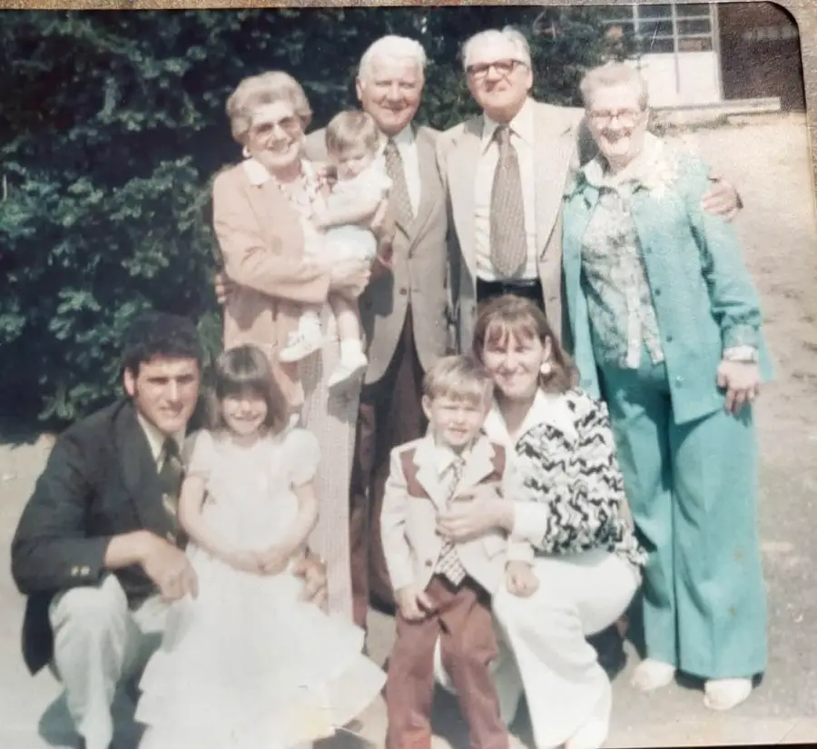 A vintage photo of three men, three women, and three children posing outdoors, with trees in the background. One woman holds a baby, and everyone is dressed in formal or semi-formal clothing, smiling at the camera.