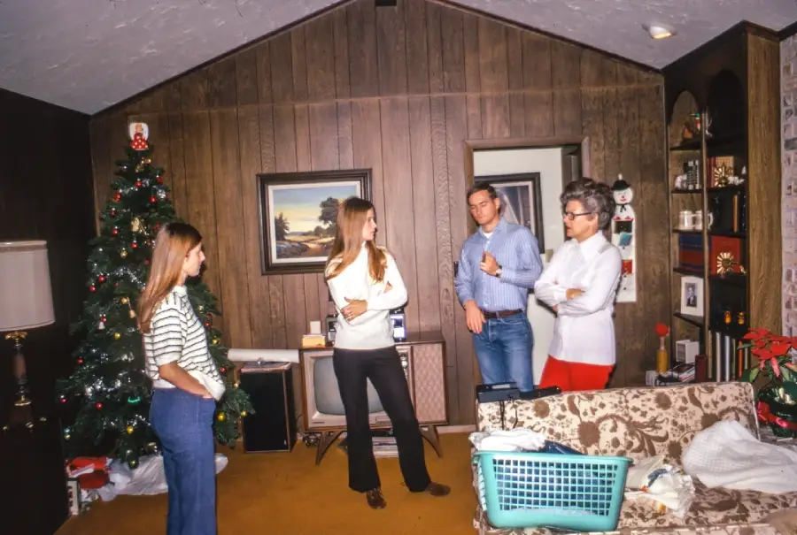 Four people stand and talk in a wood-paneled living room decorated for Christmas, with a tree, framed art, and a laundry basket on the couch in the foreground.