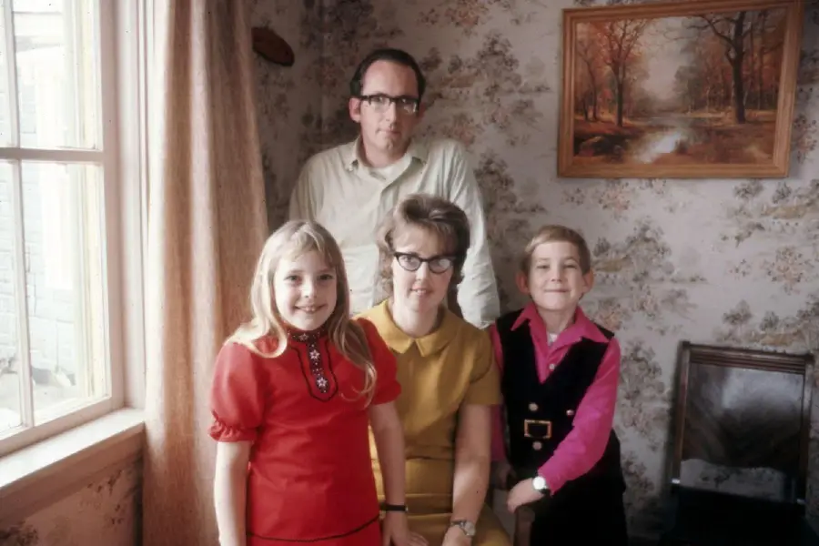 A family of four poses indoors: a man stands behind a seated woman, with a young girl and boy standing in front. The room has floral wallpaper and a window with curtains. A framed landscape painting hangs on the wall.