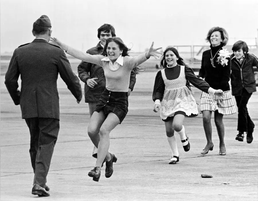 A young girl joyfully runs with open arms toward a uniformed man on a runway, followed by smiling children and a woman.